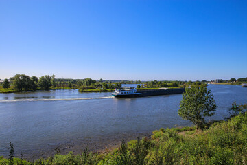 Fototapeta na wymiar View over green rural landscape on river Maas with inland waterway vessel against blue summer sky - Between Roermond and Venlo, Netherlands
