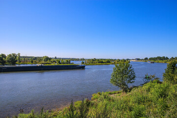 Fototapeta premium View over green rural landscape on river Maas with inland waterway vessel against blue summer sky - Between Roermond and Venlo, Netherlands