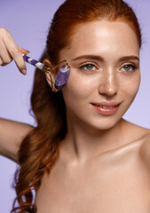 Natural beauty and skin care concept. Portrait of smiling redhead woman using quartz stone roller for gua sha massage, massaging facial skin under eyes for puffiness, purple background