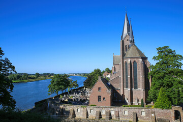 View on roman catholic church with cemetery at river Maas with lush green trees against blue summer sky - Kessel (Limburg), Netherlands