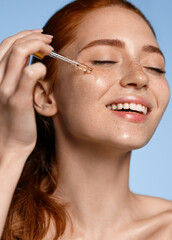 Happy smiling woman applying serum on her face, close eyes with pleasure. Redhead girl holding...