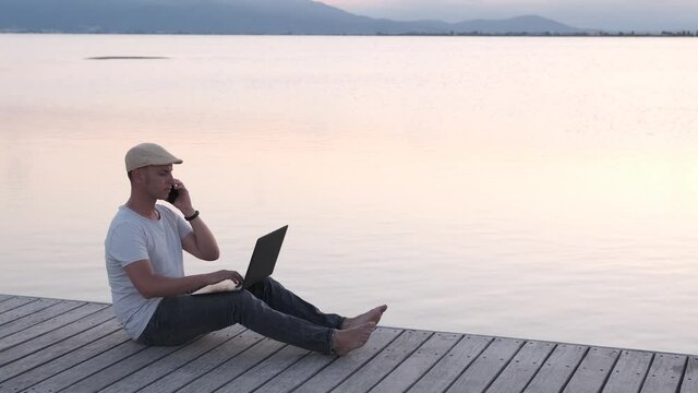 Male traveler sitting on the shore using laptop and smartphone. Digital nomad or remote worker concept