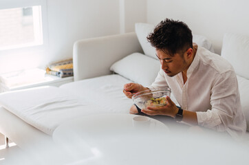 Portrait of young handsome Chinese man eating a fresh, delicious and healthy tomate salad with tune on the sofa in the living room of a minimalist and clean home