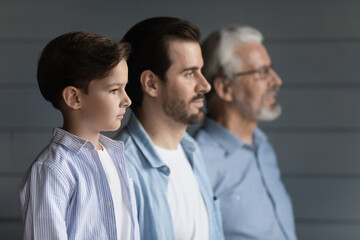 Three generations of Caucasian men stand in line pose on grey wall background together. Small teen boy child with young father and old grandfather show family unity and support. Bonding concept.