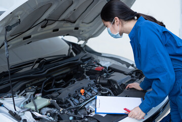 Side view, medium shot of Asian woman in a blue jumpsuit, standing in front of a car hood, looking at the engine, checking or evaluating. Female worker or engineer and car service maintenance concept.