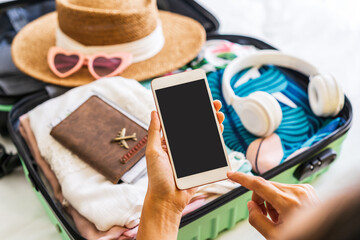 Young woman traveler sitting on the bed using smartphone and packing her suitcase preparing for travel on summer vacation