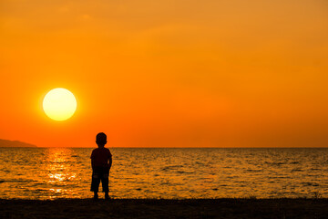 Silhouette little boy stand on sand beach looking to sea on holiday with sun sky background.