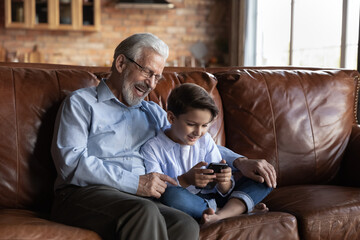 Obraz premium Loving happy old Caucasian grandfather sit relax on sofa at home with cute little grandson use smartphone together. Smiling mature granddad with small boy child rest communicate online on cellphone.