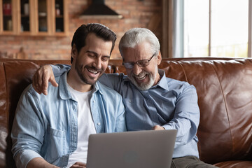 Overjoyed millennial man child relax on sofa with smiling senior father use modern laptop gadget...