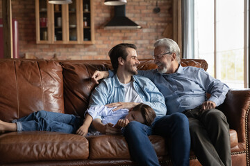 Three generations of men relax on sofa in cozy living room talk and chat on family weekend. Happy young Caucasian man rest have fun on couch at home with smiling old father and small teen son.