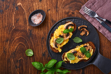 Scrambled eggs with fried mushrooms and basil on bread on black table background. Homemade breakfast or brunch meal - scrambled eggs and mushrooms sandwiches. Top view with copy space