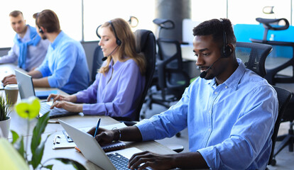 Smiling african male call-center operator with headphones sitting at modern office with collegues...
