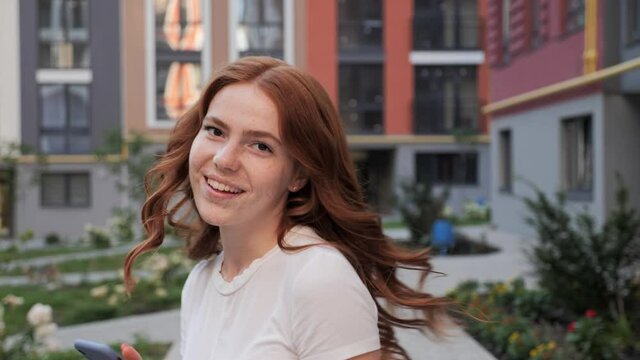 Gorgeous young Woman with Red hair is smiling Charmingly at the Camera Holding Mobile Phone. An attractive young Girl is walking down the Streets, turning to Camera, showing sincerely Emotions.