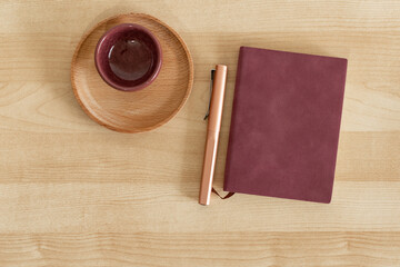 Burgundy notebook and pen, next to cup and wooden dish, on wooden desk.