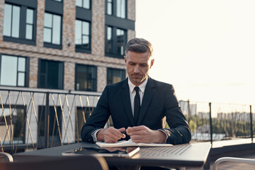 Confident mature businessman making notes while sitting on business center terrace