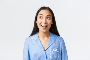 Close-up of excited beautiful asian girl hearting amazing announcement, standing in blue pajama and looking upper left corner with amazement, posing over white background thrilled