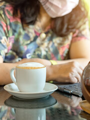 Hot Cappuccino coffee for quarantined woman wear face mask and work from home by using tablet and stress relief with hot coffee. Concepts of Covid-19 (Coronavirus disease). Selective focus on coffee.