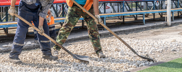 A brutal unshaven worker in overalls smooths gravel for asphalt in a treadmill stadium. Hard work with a shovel in hot weather, like in the movie