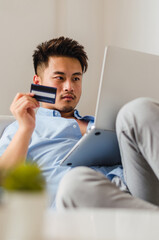 Young handsome Chinese man at home lying on the couch and shopping online with credit card and laptop