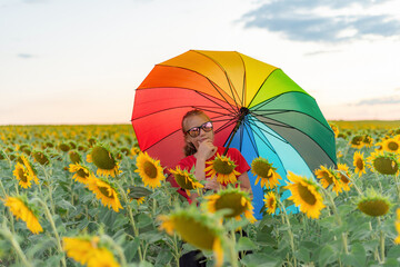 A girl in sunglasses and a colored umbrella in a field of sunflowers looks thoughtfully aside.