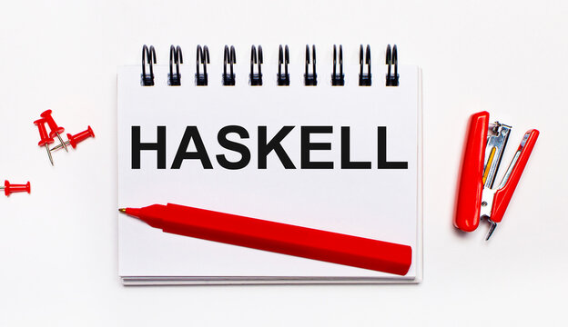 On a light background, a red pen, a red stapler, red paper clips and a notebook with the inscription HASKELL