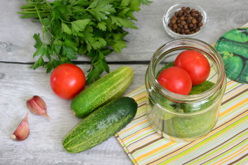 preserved vegetables in jar, pickled cucumbers and tomatoes with parsley, garlic and allspice on gray wooden background with yellow towel