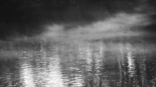 4k black and white filtered stock video footage of early sunrise morning misty landscape. Clouds of thick white fog rolling and flying over surface of calm peaceful water of dark river