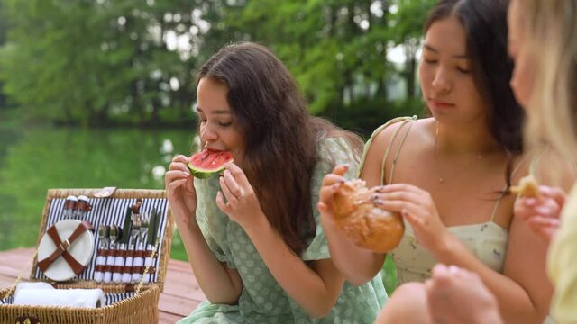 Group of friends having fun on picnic, sitting on pier eating fresh bread and watermelon. Portrait of beautiful young woman
