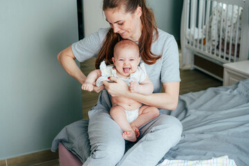 A young mother is dressing up her cheerful infant baby sitting on her knees and showing tongue.