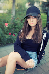 beautiful teenage lady with long legs in jeans short sits on skateboard in park setting. Her long silky hair on shoulders.