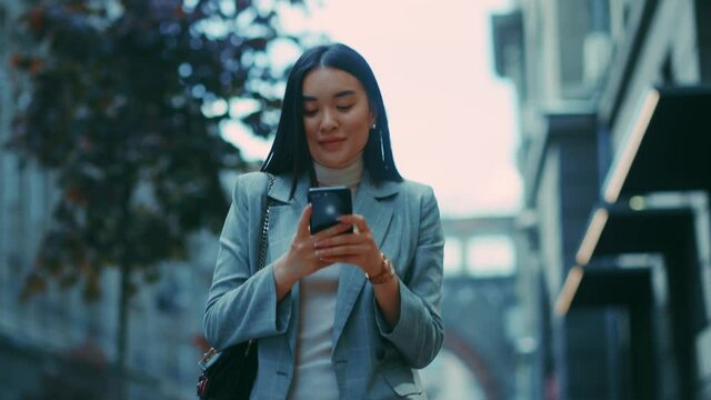 Young beautiful asian woman in business outfit outdoors. Confident corporate woman happy with good job heading to office using mobile phone.