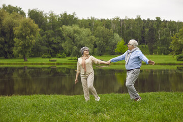 Naklejka na ściany i meble Happy loving senior couple having good time in nature. Cheerful man and woman walking in green park on fine summer day. Active old people strolling together along grassy river bank in the countryside
