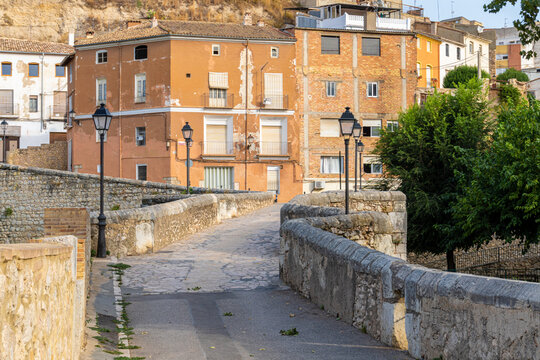 Narrow country road over an old stone bridge, with some houses in the background, on a sunny day.
