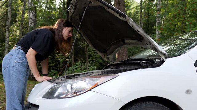 Confused young woman looks at the engine of her car. Car breakdown while traveling.