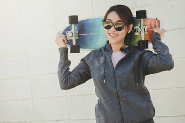 Happy Asian woman wearing sunglasses holding surfskate skateboard behide her neck against concrete wall © interstid