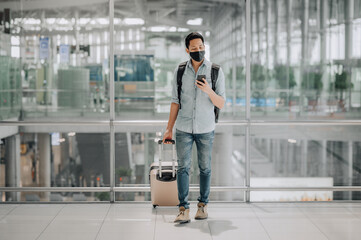 Fototapeta na wymiar Portrait of Asian man with face mask to protect from coronavirus in blue shirt with luggage using smartphone while walking