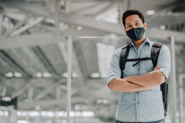 Portrait of handsome Asian man with face mask to protect from coronavirus in blue shirt standing with backpack crossed arms looking at camera 