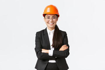 Professional smiling asian businesswoman in safety helmet and suit cross arms and looking...