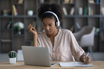 Pensive millennial African American female student in earphones study online on laptop handwrite take notes. Focused young ethnic woman in headphones work distant on computer write summarize in pad.