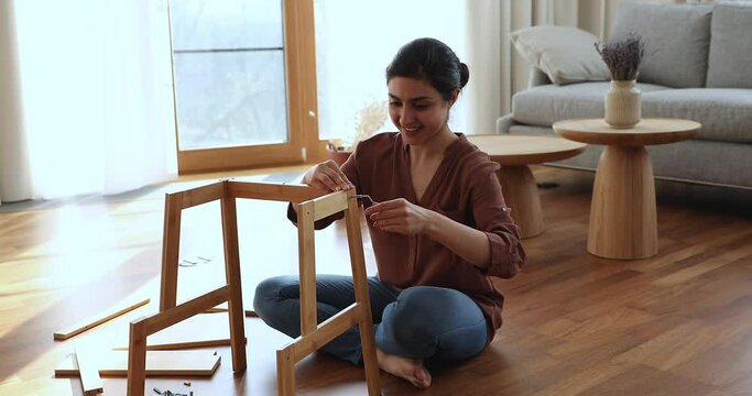 Young 25s happy Indian woman sits on floor collects assembling details of wooden furniture boards parts using hand tools screwdriver. Enjoy new purchase, modern furniture shop, do-it-yourself concept