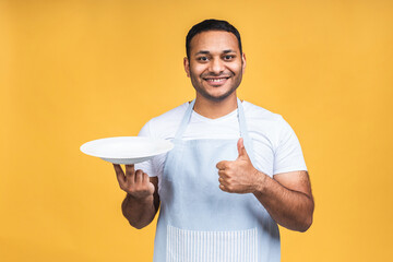 Indian african american chef cook or baker man in striped apron isolated over yellow background. Cooking food concept. Mock up copy space. Hold empty blank plate with place for food. - 451048363