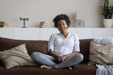 Smiling millennial African American woman sit rest on couch in living room watch TV on weekend break. Happy young ethnic female renter tenant relax on sofa at home enjoy television program online.