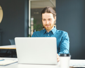 Happy young male office employee in shirt sitting at desk in front of laptop computer and looking at screen with smile, enjoying work in office. Cheerful man using digital technologies at workplace