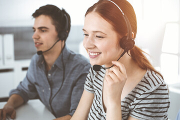 Casual dressed young woman using headset and computer while talking with customers online. Group of operators at work in sunny office. Call center