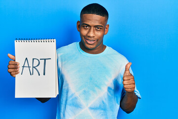 Young black man holding art notebook smiling happy and positive, thumb up doing excellent and approval sign