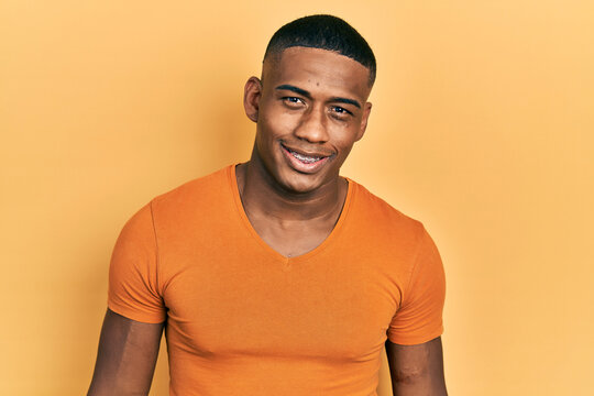 Young Black Man Wearing Casual Orange T Shirt Looking Positive And Happy Standing And Smiling With A Confident Smile Showing Teeth