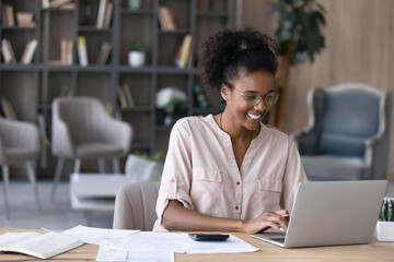 Happy millennial African American woman manage household budget calculate expenses expenditures on machine. Smiling young biracial female pay bills taxes online on computer. Finances concept. - obrazy, fototapety, plakaty