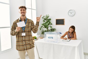 Young handsome man voting putting envelop in ballot box smiling happy pointing with hand and finger to the side