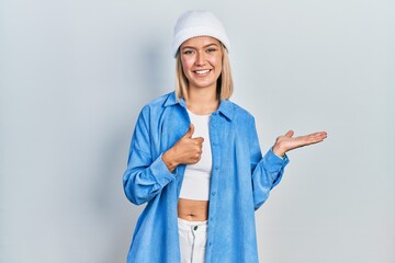 Beautiful blonde woman wearing wool hat showing palm hand and doing ok gesture with thumbs up, smiling happy and cheerful