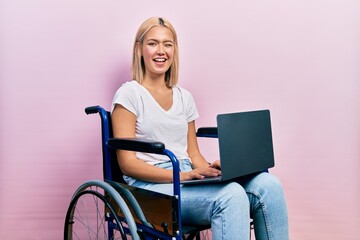 Fototapeta na wymiar Beautiful blonde woman sitting on wheelchair holding laptop winking looking at the camera with sexy expression, cheerful and happy face.